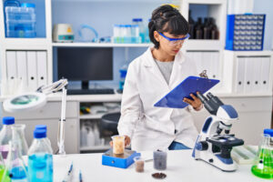 A female scientist in a lab coat inspects notes beside a microscope in a laboratory.