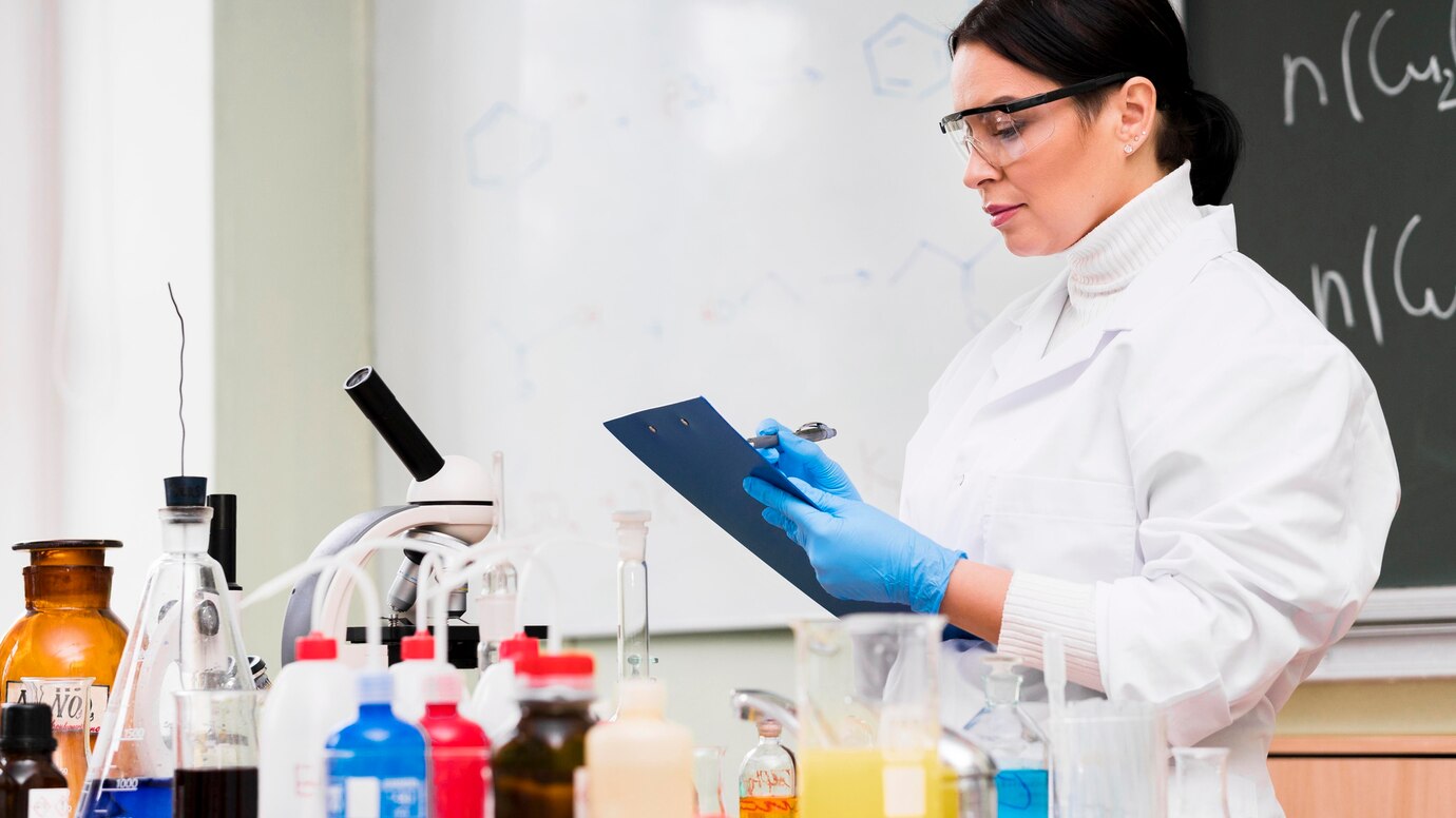 Female scientist in lab coat examining notes in a chemistry lab surrounded by various scientific equipment and chemicals.