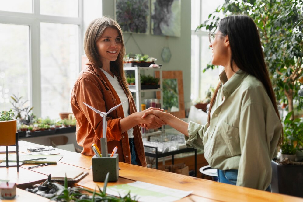 Two women shaking hands in a bright office filled with plants, one holding a model wind turbine.
