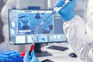 Scientist in a lab coat using a pipette to transfer red liquid into a test tube, with computer screens in the background.