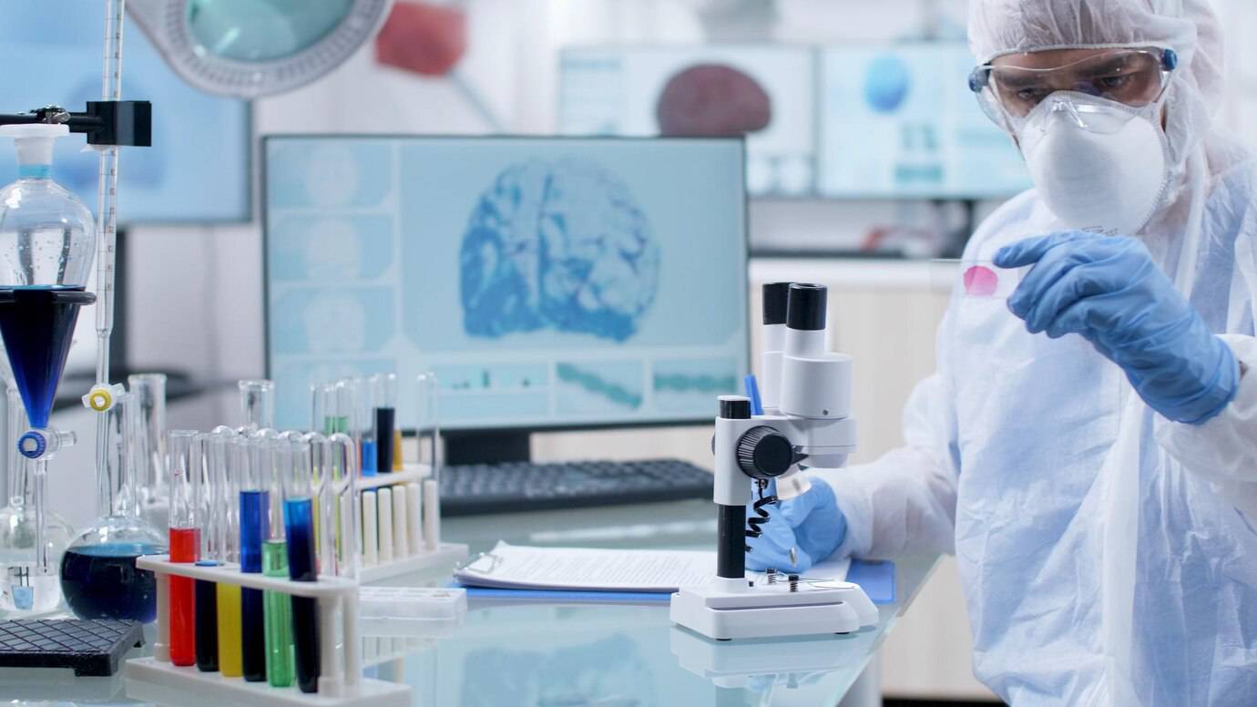 Scientist in protective gear examines a sample in a modern laboratory with a microscope and colorful test tubes.