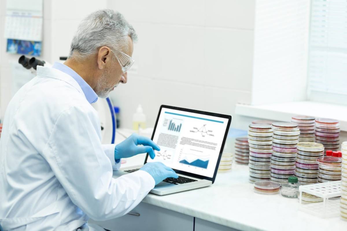 A lab technician in a white coat and gloves analyzes data on a laptop, surrounded by stacks of petri dishes 