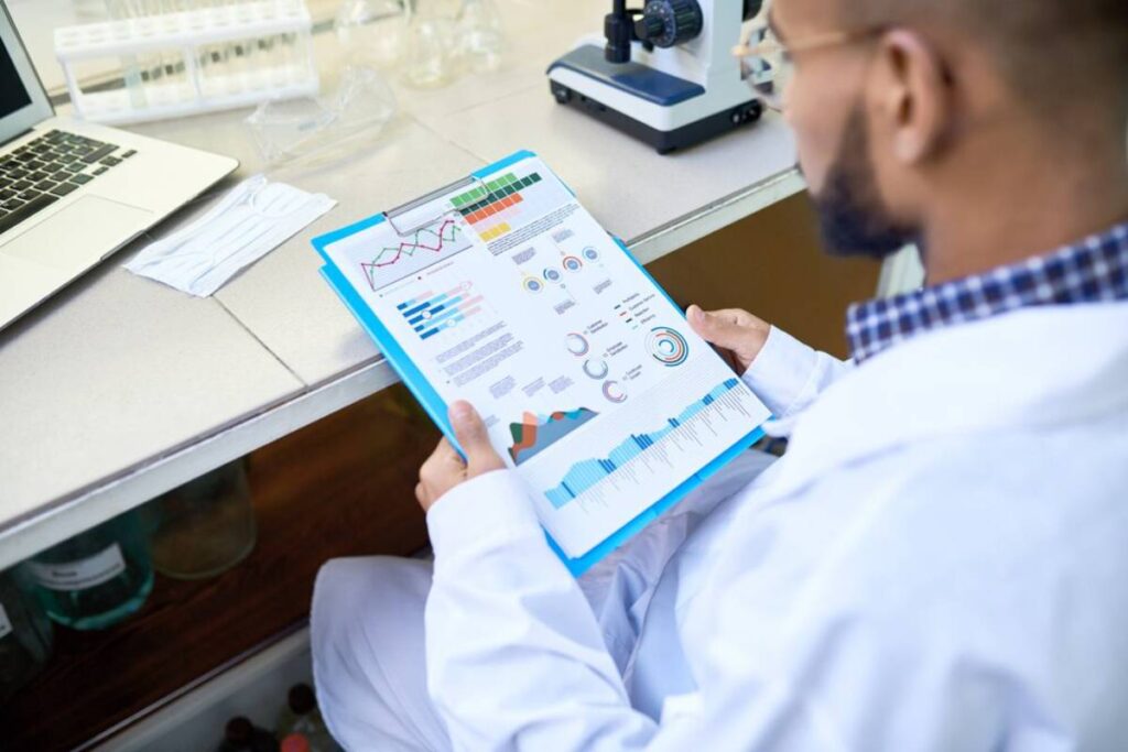 A researcher in a lab coat examines colorful graphs and charts on a document, with a microscope and laptop in the background.