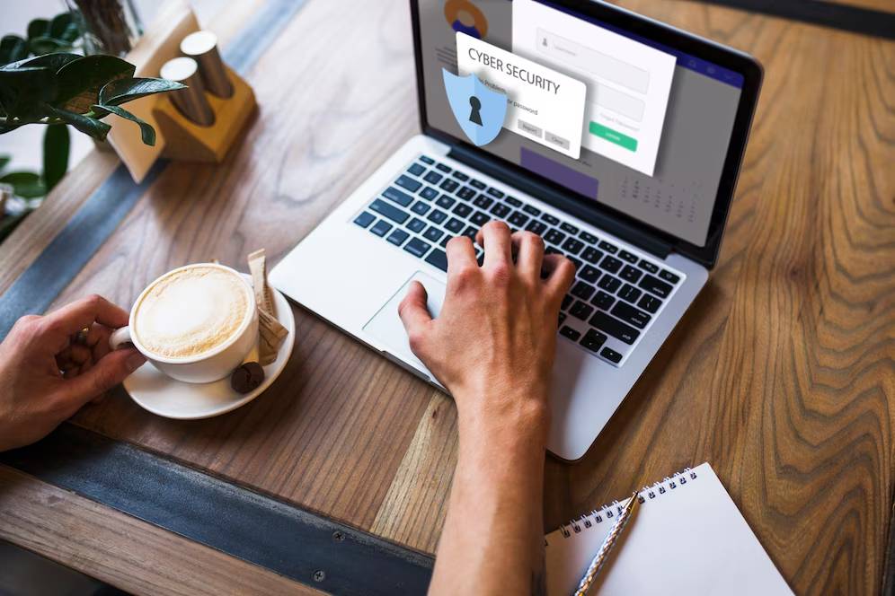 Person working on a laptop showing a cyber security screen, with a cup of coffee and notepad on a wooden table.