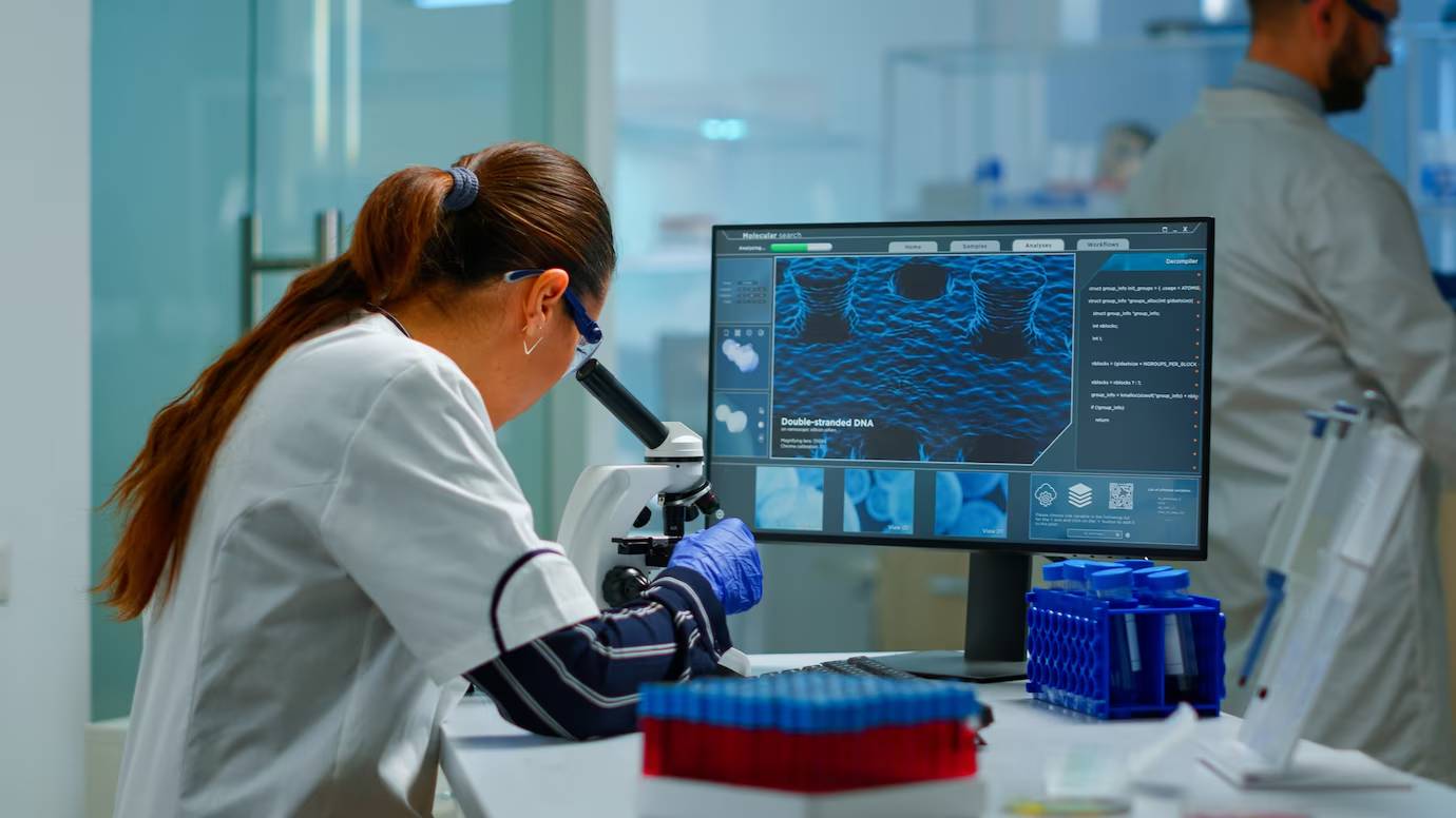 Scientist using a microscope in a lab, with a digital screen displaying DNA images in the background.