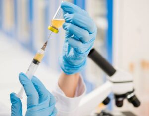A gloved hand holds a syringe filled with yellow liquid, drawing from a vial, with a microscope visible in the background.