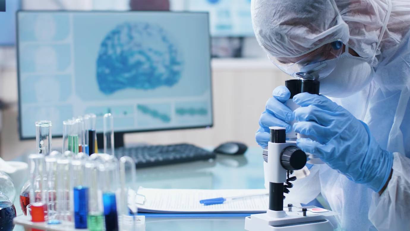 Scientist in protective gear using a microscope in a laboratory with test tubes and a computer displaying graphs.