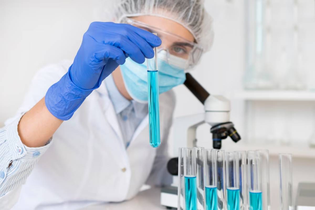 Scientist in lab coat and gloves examines a blue liquid in a test tube, with microscope in background.