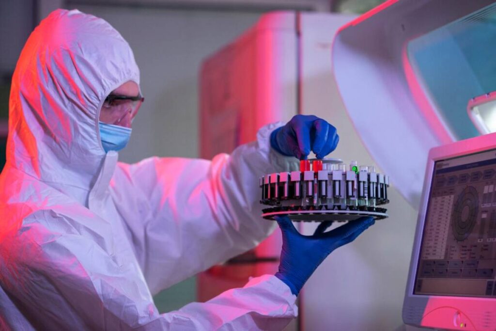 Person in protective gear holding a tray of test tubes in a laboratory setting.
