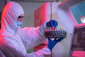 Person in protective gear holding a tray of test tubes in a laboratory setting.