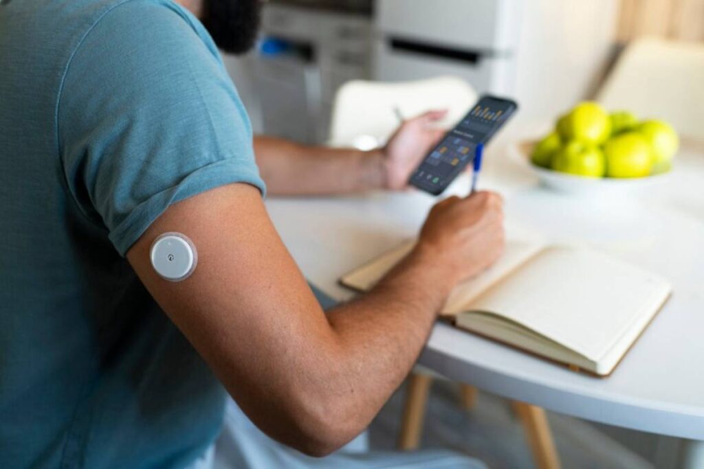 Man checking smartphone while wearing a glucose monitor on his arm, sitting at a kitchen table.