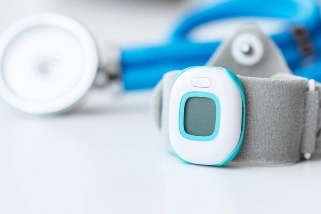 A close-up view of a digital fingertip pulse oximeter attached to a grey strap with medical equipment in the background.