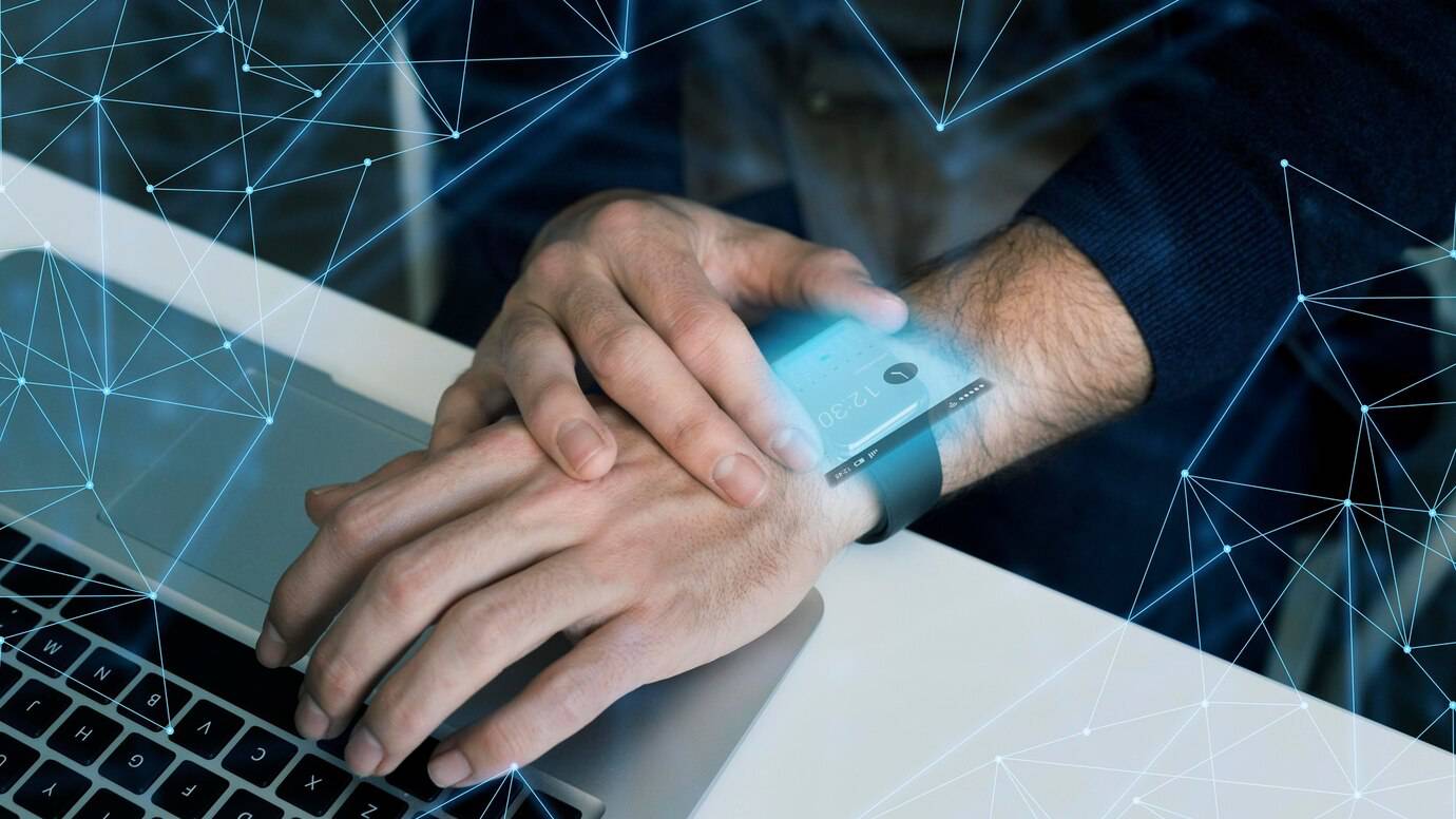A person using a futuristic device on their wrist while working on a laptop overlaid with digital connections.