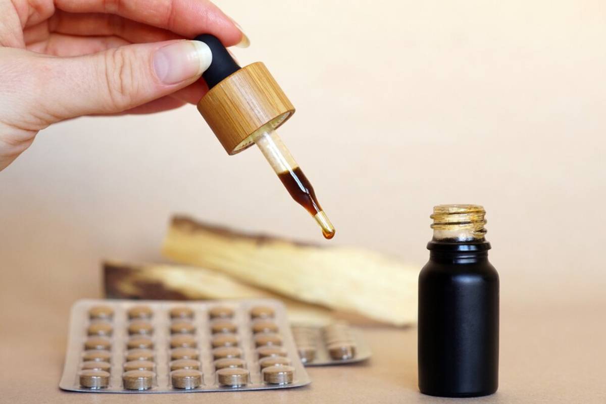 A hand holds a dropper dispensing liquid from a small bottle, with herbal supplements in the background.