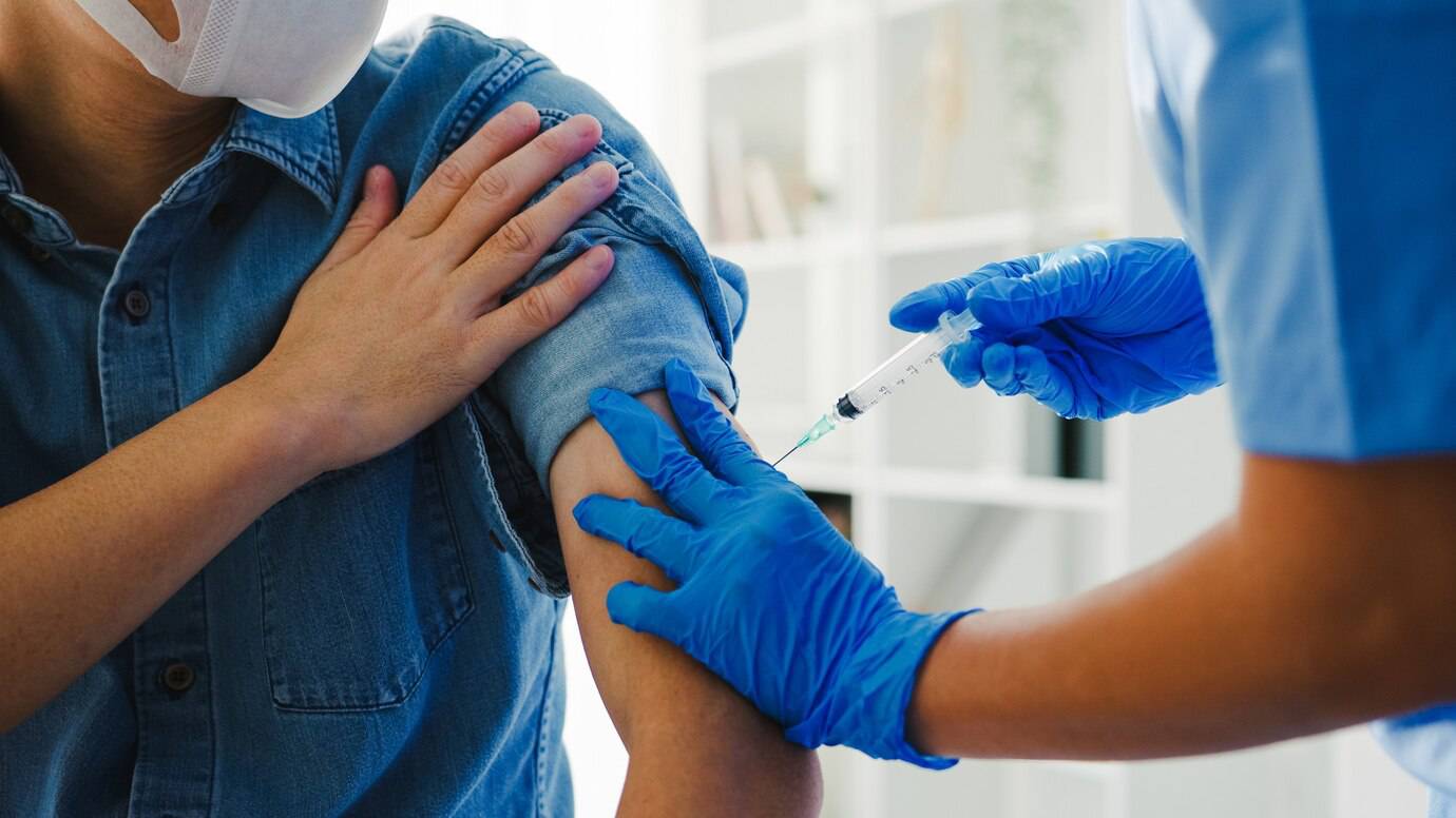 Healthcare professional in blue gloves administering a vaccine to the arm of a person wearing a mask and denim shirt.