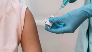 Healthcare worker in gloves drawing vaccine from a vial into a syringe next to a patient's arm.