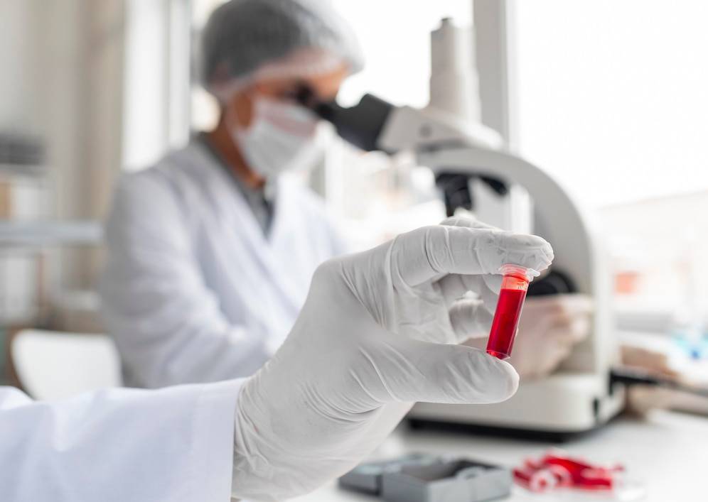 Scientist in a lab examining a red substance in a test tube, with a microscope in the background.