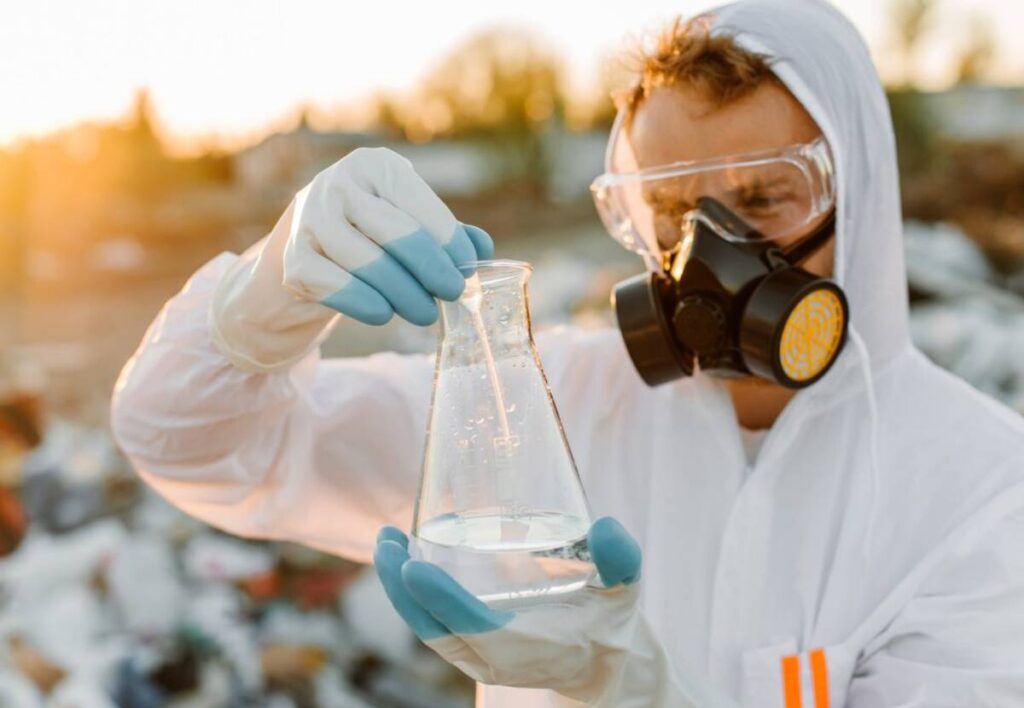 Scientist in protective gear examines a flask of clear liquid at a landfill site during sunset.