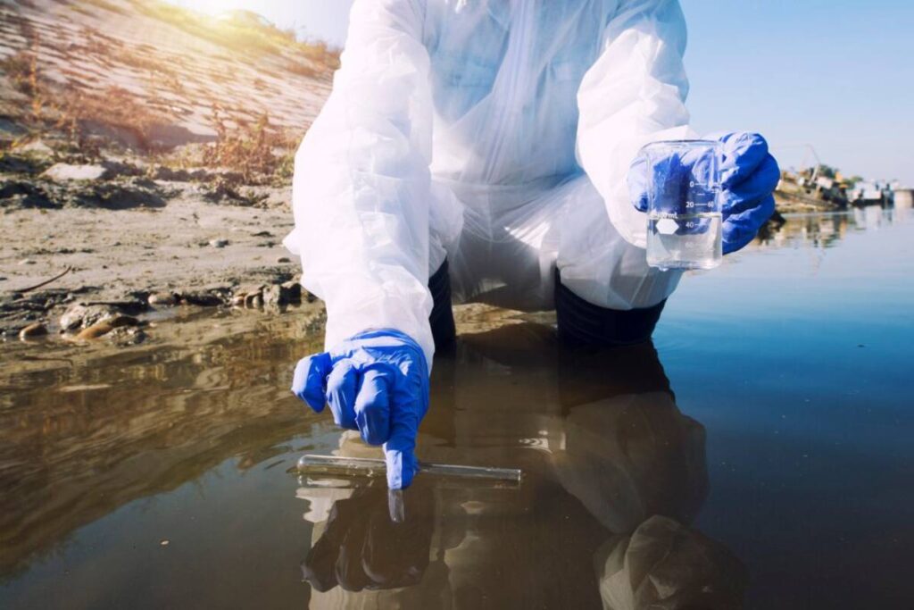 Scientist in protective suit collecting water samples from a polluted river for environmental testing.