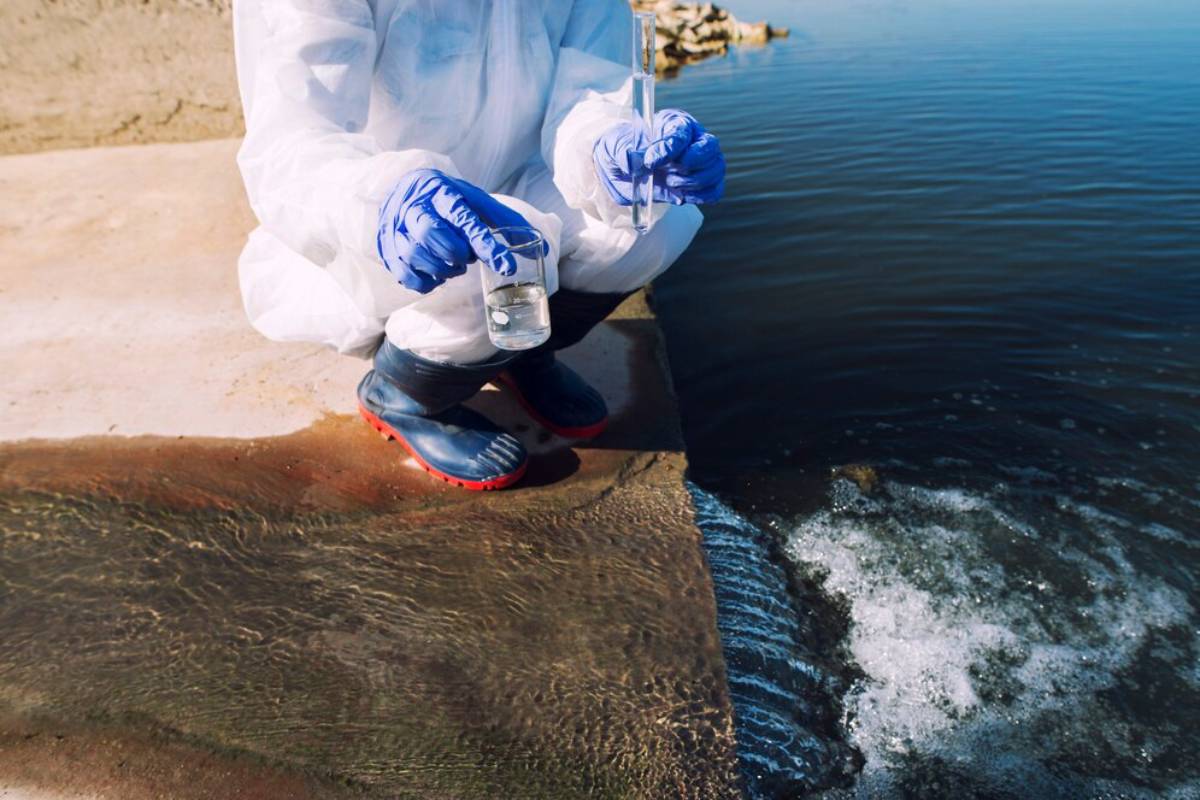 A person in protective gear collecting water samples from a river edge.