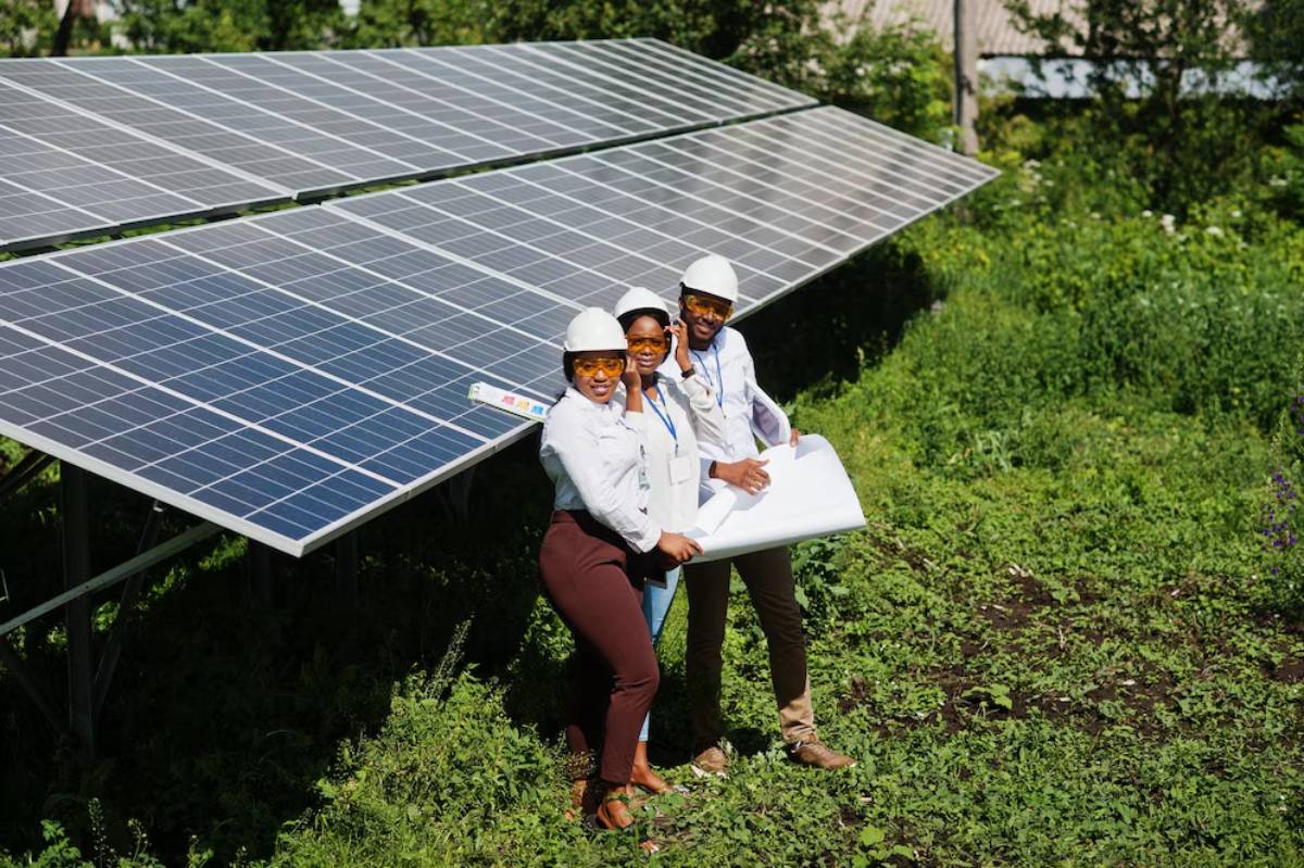Three engineers in hard hats and safety glasses examine a solar panel installation in a lush, green field.