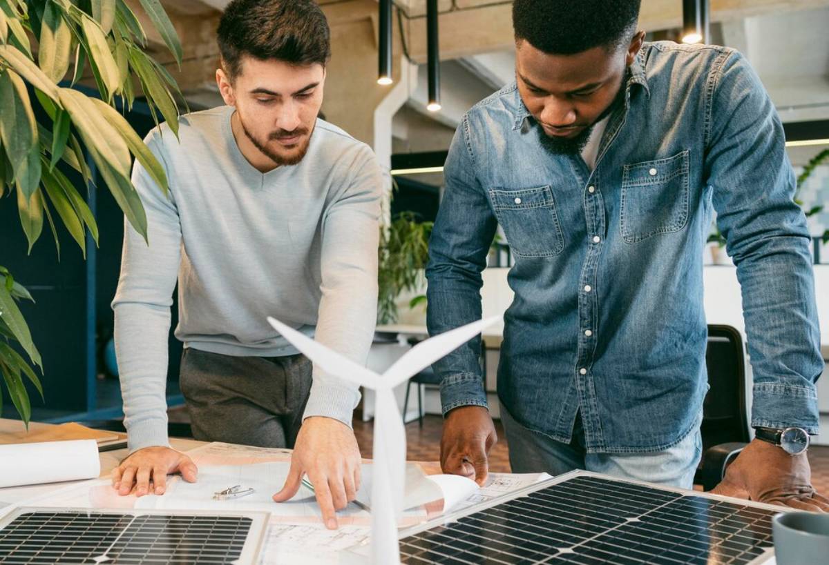 Two men examining renewable energy plans with a small wind turbine and solar panel on a table.